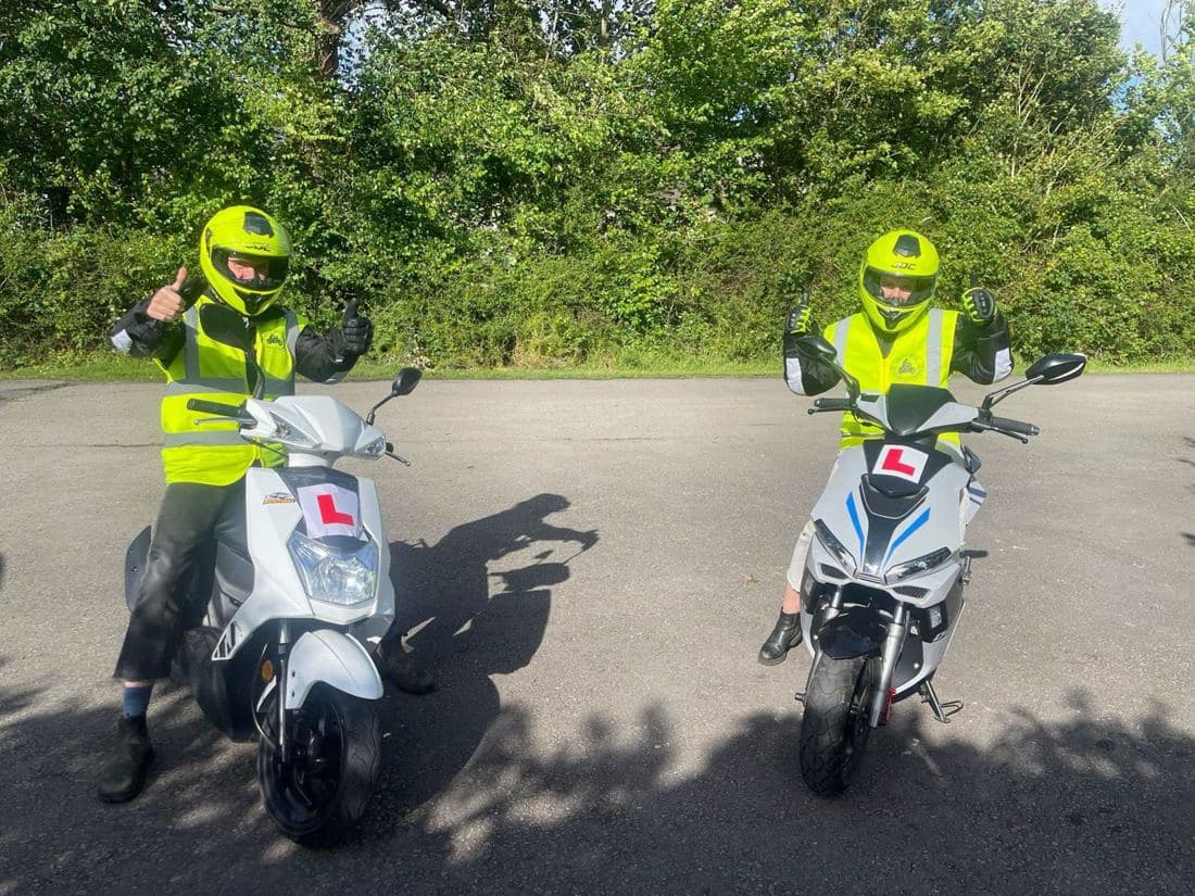 Two learner riders in hi-vis giving a thumbs up on white L-plated mopeds during CBT motorcycle training
