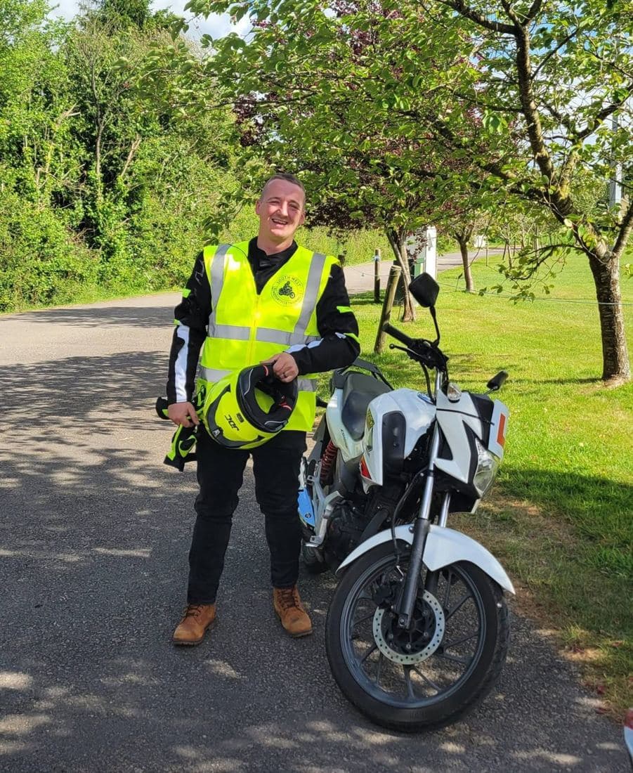 A rider in a South West Motorcycle Training hi-vis holding his helmet beside a white 125cc training motorcycle