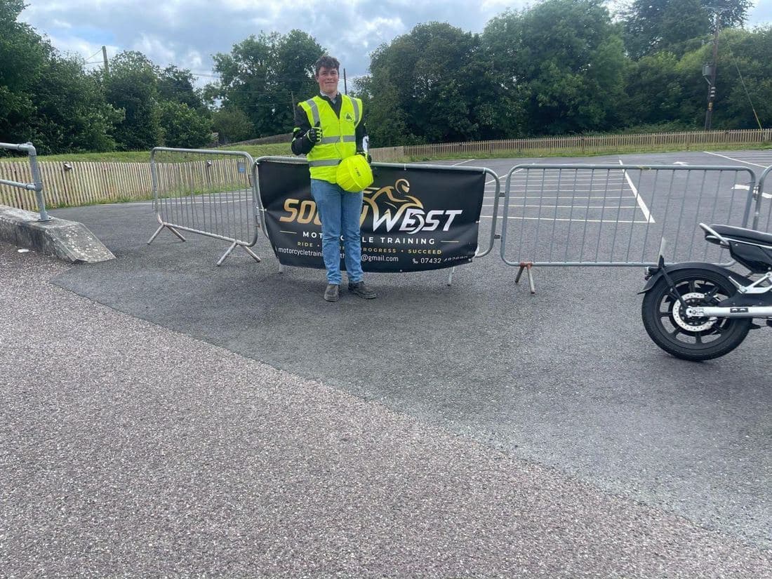 A learner rider in hi-vis with a yellow helmet in front of the South West Motorcycle Training banner on the training pad