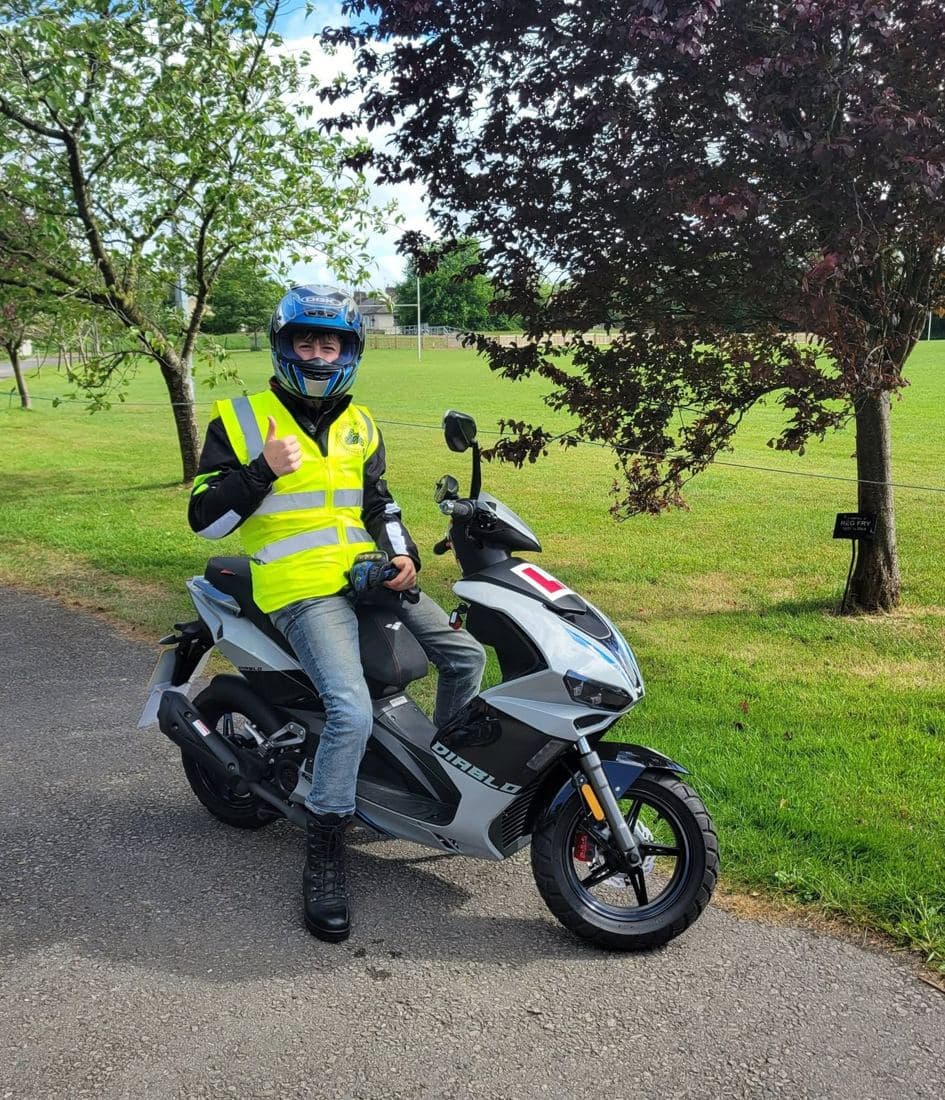 A young learner rider in hi-vis and a blue helmet sitting on an L-plated scooter, giving a thumbs up A young learner rider in hi-vis and a blue helmet sitting on an L-plated scooter, giving a thumbs up