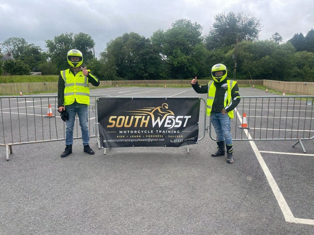 Two riders in helmets and hi-vis giving a thumbs up either side of the South West Motorcycle Training banner