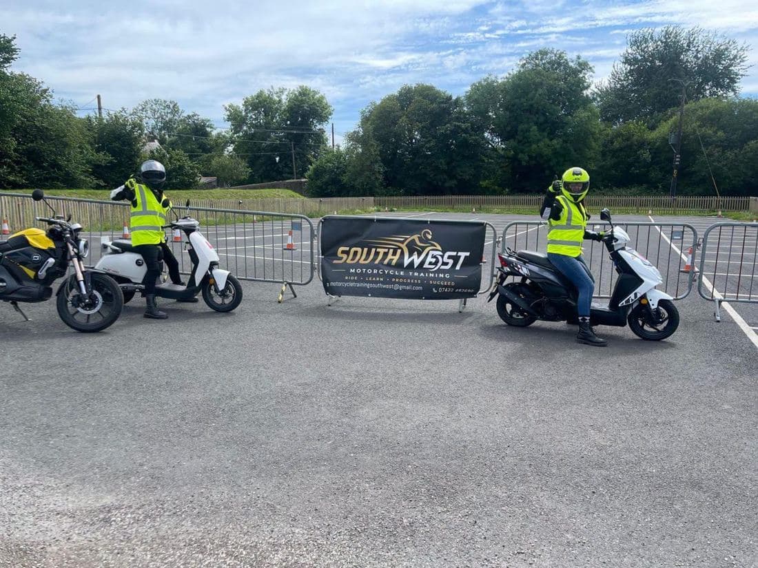 Two riders in hi-vis beside L-plated scooters on the manoeuvring pad, with the South West Motorcycle Training banner behind them